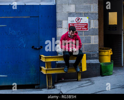 Mann sitzt auf Metall Barriere, über Smartphone, Zigarette, Central London, England, Großbritannien Stockfoto