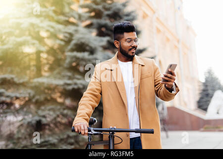 Schöne bärtige Geschäftsmann im klassischen Anzug ist mit einem Smart Phone und lächelnd beim Reiten Fahrrad in der Stadt Stockfoto