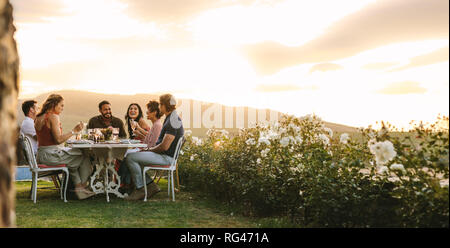 Gruppe von jungen Freunden heraus hängen mit Getränken zum Abendessen im Freien. Junge Männer und Frauen sitzen um einen Tisch mit Essen und Getränken. Stockfoto