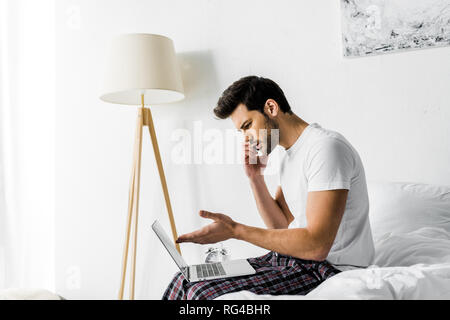 Mann auf Smartphone und mit Laptop auf dem Bett besorgt Stockfoto