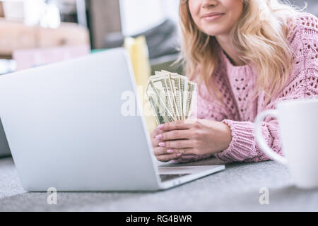 7/8-Ansicht fröhliche Frau mit Dollar Banknoten, während in der Nähe von Laptop liegen Stockfoto