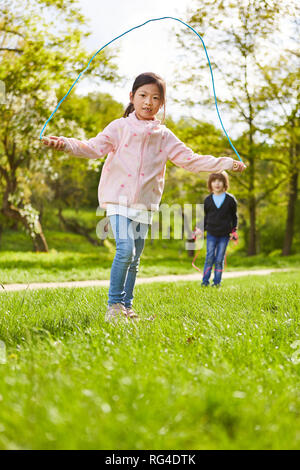 Asiatische Mädchen im Seil springen auf einer Wiese mit Freundin im Hintergrund Stockfoto