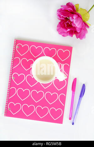 Spirale Notebook mit rosa und weißen Herzen Design. Tasse Tee in Weiß Becher, pink silk Blume und rosa und lila Stifte auf weißem Hintergrund in Flach Stockfoto