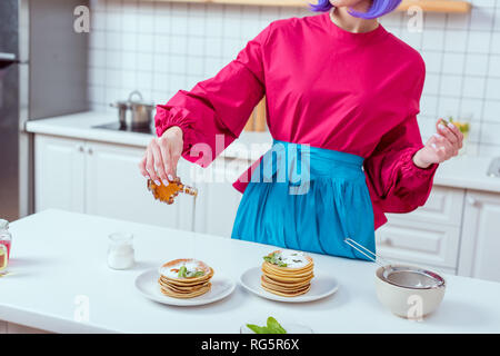 7/8-Ansicht von Hausfrau in bunten Kleidern gießen Sirup auf Pfannkuchen in der Küche Stockfoto