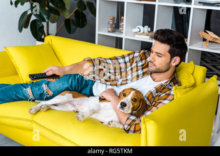 Stattlicher Mann mit Fernbedienung Fernsehen und liegen auf einem Sofa mit Beagle Hund Stockfoto