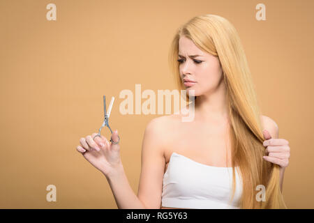 Verärgert junge Frau mit langen blonden Haaren holding Schere isoliert auf beige Stockfoto