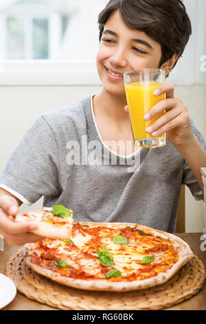 Portrait Of Happy Teen Boy mit Genuss essen Pizza und trinken Orangensaft, fröhlichen Jugendlichen Freizeitgestaltung in der Pizzeria, lecker, aber un Stockfoto