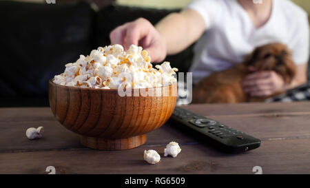 Hölzerne Schüssel mit gesalzenem Popcorn und TV-Fernbedienung auf hölzernen Tisch. Im Hintergrund, ein Mann mit einem roten Hund Fernsehen auf der Couch. Stockfoto