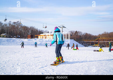 Ein junges Mädchen in einer speziellen farbigen Anzug für Winter Sport und Spaß lernt ein snowboard an einem sonnigen Tag zu reiten. Stockfoto