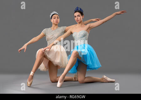 Adorable Balletttänzer sitzen auf Knien, eine Hand heben und Kamera im Studio. Ballerinas in Kleider und Ballett Schuhe auf grauem Hintergrund posiert. Konzept der Leistung und Ballett. Stockfoto