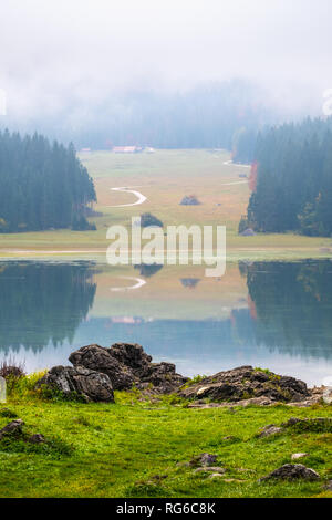 Blick auf den Laghi di Fusine an einem nebligen Morgen mit einigen Felsen am Strand und im Gebirge Mangart in der Nähe von Tarvisio in Italien Stockfoto