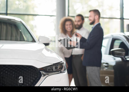 In der Nähe von weißen Auto Scheinwerfer. Glückliches Paar in der Nähe von Autohaus. über den Kauf des Fahrzeugs. Manager in der dunklen blauen Jacke, Schlüssel und schwarzen Ordner, Dokumente. Stockfoto