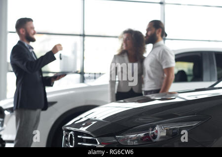 Schwarzes Auto Motorhaube schließen. Manager gegenüber Kunden des Autohauses. Autohändler holding Autoschlüssel. Paar Einkauf neue teure Autos in der modernen Showroom. Stockfoto
