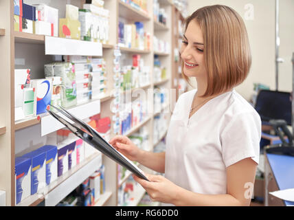 Happy Apotheker in der Nähe von Regalen mit Medikamenten in der Apotheke. Schöne Frau in weißer uniform Lächeln, Ordner in den Händen. Spezialist in der pharmazeutischen Industrie zu arbeiten. Stockfoto