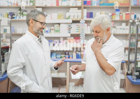 Älterer mann Consulting mit Apotheker über die Wahl der Medikamente. Chemiker in Gläsern und weiße Uniform Lächeln, Rentner. Alte Kunden denken, holding Blisterverpackung. Stockfoto