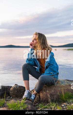 Finnland, Lappland, junge Frau sitzt auf einem Felsen am See Stockfoto