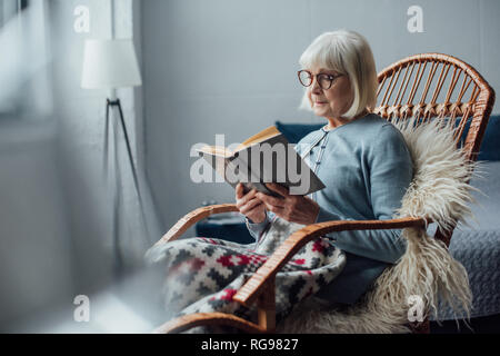Ältere Frau in Gläsern sitzen in Rattan Schaukelstuhl und lesen Buch zu Hause Stockfoto