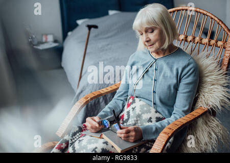 Nachdenklich senior Frau sitzt in Rattan Schaukelstuhl mit Buch zu Hause Stockfoto