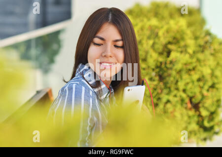 Ein junges Mädchen der asiatischen Aussehen sitzt auf einer Bank und hört Musik in der Nähe des grünen Büschen Stockfoto