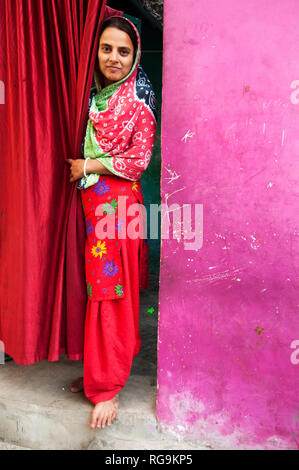 Indien. Bihar. Hohenstadt. purani Gudri (alte Slum). Eine junge Frau tragen bunte shawar kameez blickt von einem roten Vorhang Tür. Stockfoto