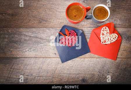 Festliche Komposition auf rustikalen Holztisch, rote und graue Tasse mit Marshmallows oder Kakao, Herz in Umschlägen, getrocknete rosa Blüten. Stockfoto