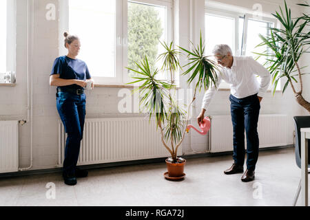 Manager Bewässerung von Pflanzen im Aufenthaltsraum, während Arbeiter trinkt Kaffee Stockfoto