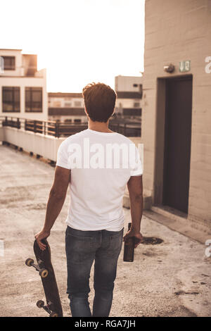 Rückansicht des jungen Mannes mit Skateboard und Bier Flasche auf der Dachterrasse zu Abend dämmerung Stockfoto