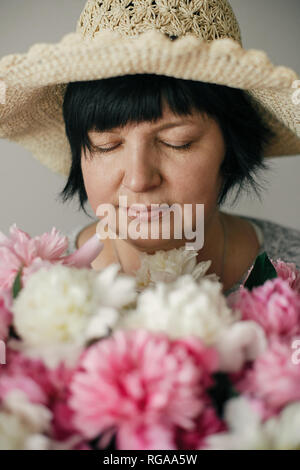 Elder schöne Frau in hat mit vielen Pfingstrose Blumen posieren. Glückliche Mutter mit großen Blumenstrauß aus rosa Pfingstrosen von Kindern. Glückliche Mütter Tag Konzept. Ich Stockfoto