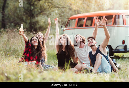 Eine Gruppe junger Freunde mit Getränken sitzen auf Gras vor der retro Minivan auf einem Roadtrip durch die Landschaft. Stockfoto