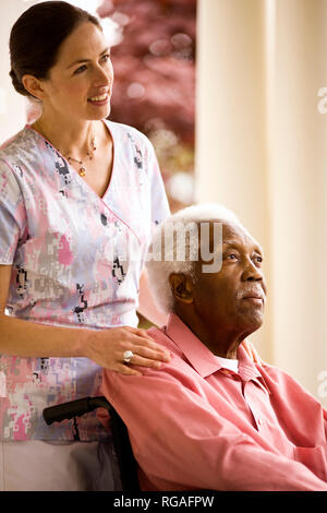 Lächelnd Krankenschwester setzt unterstützende Hand auf die Schultern ein älterer Mann, als er in einem Rollstuhl auf einer Veranda sitzt. Stockfoto