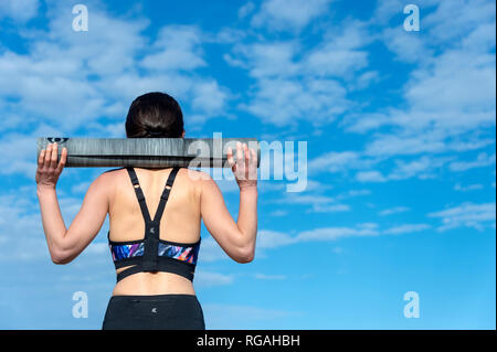 Frau Sportswear eine Yogamatte über ihre Schultern außerhalb tragen in der Sonne mit blauem Himmel Stockfoto