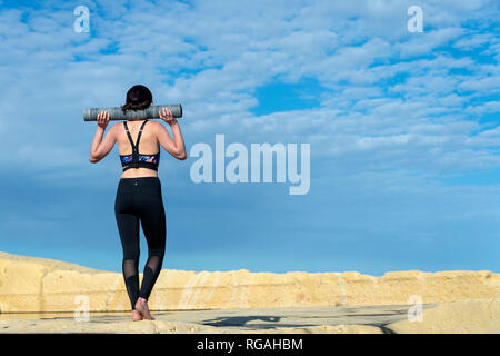 Frau Sportswear eine Yogamatte über ihre Schultern außerhalb tragen in der Sonne mit blauem Himmel Stockfoto