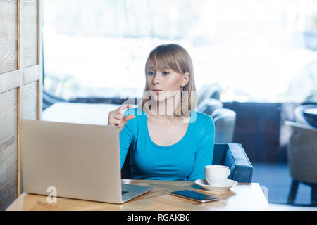 Wenig! Portrait von attraktiven jungen Mädchen mit blondem Haar in blauer Bluse Sitzen im Cafe, Arbeiten und Videokonferenz auf Laptop, er freue Stockfoto