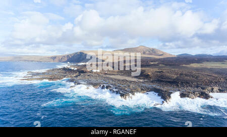 Luftaufnahme von einem Fischerdorf auf einem felsigen Ufer durch hohe Wellen geschlagen, mit vulkanischen Berge am Horizont,, Lanzarote, Kanarische Inseln, Spanien. Stockfoto