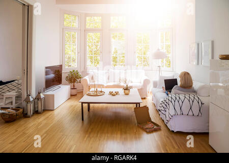 Rückansicht der Blonde reife Frau sitzt auf der Couch im Wohnzimmer mit Laptop Stockfoto