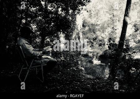 Ein junger Mann liest ein Buch neben einem Fluss, Bush Camp an der Ausgrabungen, Crediton State Forest, Queensland, Australien Stockfoto