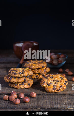Avena cookies mit Schokoladenstückchen und Haselnüsse auf einem Holztisch mit einer Schüssel mit avena Flocken und einem Löffel, ein weiterer Haselnüsse mit Schale Stockfoto