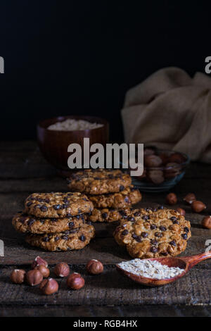 Avena cookies mit Schokoladenstückchen und Haselnüsse auf einem Holztisch mit einer Schüssel mit avena Flocken und Haselnüsse mit Schale und einem Tuch Stockfoto