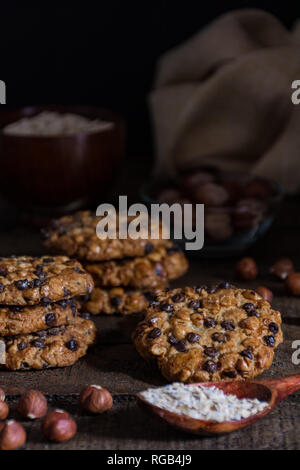 Avena cookies mit Schokoladenstückchen und Haselnüsse auf einem Holztisch mit einer Schüssel mit avena Flocken und Haselnüsse mit Schale und einem Tuch Stockfoto