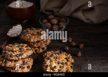 Avena cookies mit Schokoladenstückchen und Haselnüsse auf einem Holztisch mit einer Schüssel mit avena Flocken und Haselnüsse mit Schale und einem Tuch Stockfoto