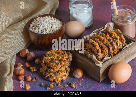 Box mit avena Cookies mit Haselnüssen, Schokolade, Chips, eine Schüssel mit avena Flocken, Gläser mit Schokolade und Mehl, Haselnüsse und zwei Ei Stockfoto