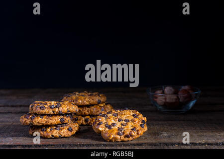 Einige avena Cookies mit Haselnüssen und Chocolate Chips auf einem alten Holztisch, eine Glasschale mit Haselnüssen und dunklen Hintergrund Stockfoto
