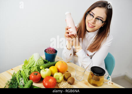 Junge süße Mädchen mit Brille, Joghurt oder Kefir in einer Flasche auf dem Hintergrund einer Tabelle mit viel Gemüse Stockfoto