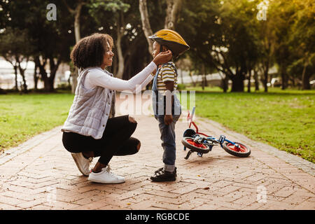 Junge Frau, Helm auf nette Junge, mit Zyklus auf der Rückseite im Park liegen. Mutter legt ihr Sohn Schutzhelm für das Reiten Fahrrad. Stockfoto