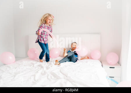 Group Portrait von zwei Glückliche weiß Kaukasischen cute adorable lustig Kinder springen auf dem Bett im Schlafzimmer. Junge und Mädchen kämpfen mit Ballons und Spaß haben Stockfoto