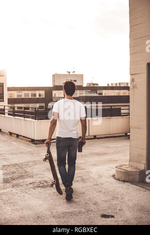 Rückansicht des jungen Mannes mit Skateboard und Bier Flasche auf der Dachterrasse zu Abend dämmerung Stockfoto