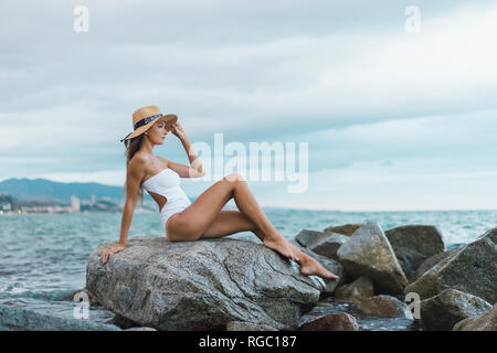 Schöne junge Frau das Tragen von Badebekleidung und Hut sitzt auf Felsen im Meer Stockfoto