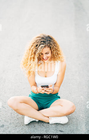 Lächelnde junge Frau sitzt auf der Straße am Handy suchen Stockfoto