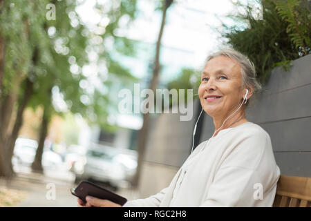 Lächelnd senior Frau sitzt auf einer Bank im Freien mit Handy und Kopfhörer. Stockfoto
