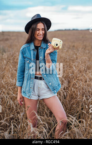 Portrait von Lachende junge Frau mit Hut und Jeansjacke in einem Maisfeld mit Kamera Stockfoto